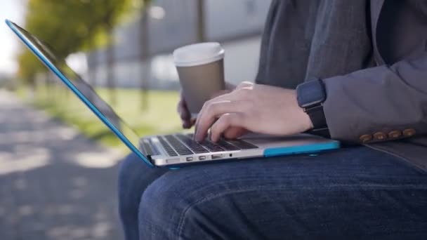 Schöner kaukasischer junger Mann in Jacke und Schal, auf der Bank sitzend, mit Laptop und Kaffeetext auf der Tastatur. Außenbereich — Stockvideo