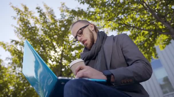 Zbliżenie: przystojny Kaukaska młodego człowieka z brodą, siedząc w ławce z laptopa. Przystojny student mężczyzna siedzi na ławce w parku i wpisując tekst na laptopie. Na zewnątrz — Wideo stockowe