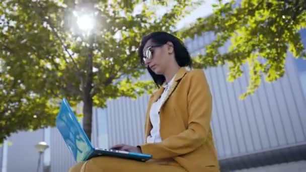 Красива ділова жінка в сонцезахисних окулярах, одягнена в костюм за допомогою ноутбука під час перерви на відкритому повітрі, молода жінка сидить на лавці в парку біля офісу бізнес-центру — стокове відео