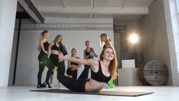 Молода усміхнена жінка розігріває дупу, розтягуючи спину, тримаючи ноги і працюючи в спортивному класі йоги. Привабливі молоді спортивні дівчата роблять йогу разом на фоні — стокове відео