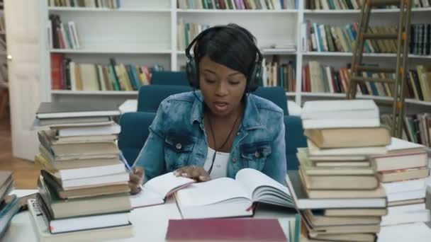 Jovem africana atraente ouvindo música no fone de ouvido enquanto faz alguma nota do livro de notas para a universidade . — Vídeo de Stock