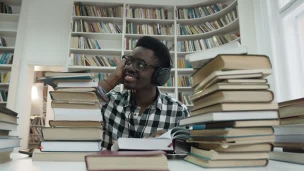 Estudiante africano sonriente escuchando música en auriculares mientras lee libro . — Vídeo de stock