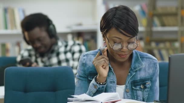 Ελκυστική αφρικανική φοιτήτριας ανάγνωση βιβλίων στη σύγχρονη βιβλιοθήκη ενώ αφρικανική αρσενικό φοιτητής χρησιμοποιώντας έξυπνο τηλέφωνο στο φόντο. — Αρχείο Βίντεο