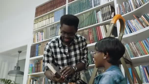 Africano chico de pie en escalera y dando libros a africano chica en universidad biblioteca . — Vídeo de stock