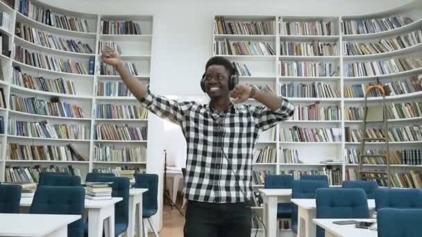 Happy african guy listening music in the headphone and dancing in the modern library. — Stock Video