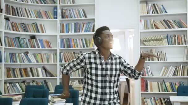Счастливый молодой африканский мужчина слушает музыку в наушниках и танцует в библиотеке . — стоковое видео