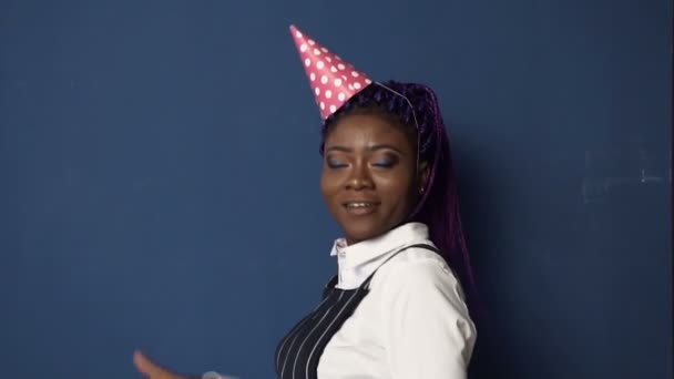 Dolly toma de niña africana sonriente en sombrero de cumpleaños bailando sobre el fondo azul . — Vídeos de Stock