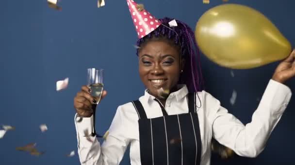 Jovem mulher africana feliz em chapéu de aniversário com vidro de champanage em uma mão e balão em outra dança enquanto confete voando para baixo no fundo azul . — Vídeo de Stock