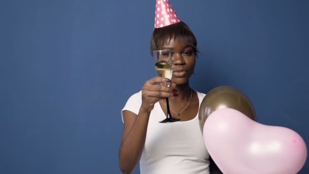 Afrikaanse meisje in hoed met ballonnen in de hand rammelende glas naar camera op de blauwe achtergrond. — Stockvideo