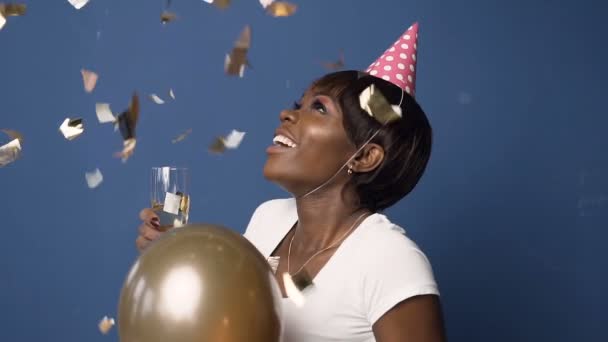 Chica africana feliz en sombrero con copa de champán en una mano y globo en otra, mirando al confeti que vuela sobre el fondo azul . — Vídeo de stock