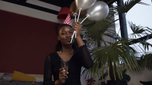 Atractiva chica africana en sombrero con copa de champanaje en una mano y globos en otra mano sentada en el suelo en la habitación roja . — Vídeo de stock