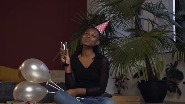Mulher jovem africana de chapéu com vidro de champanage e balões sentados no chão e possuindo na câmera na sala vermelha . — Vídeo de Stock