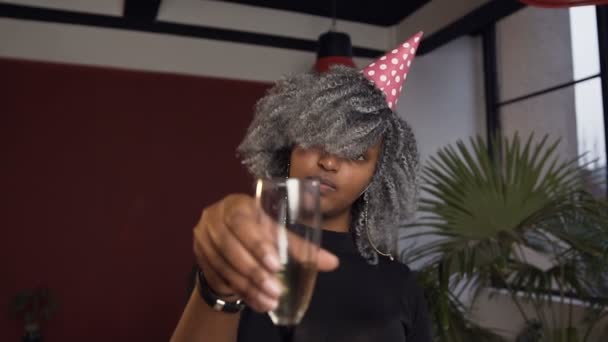 Mujer africana joven en sombrero tintineo champán de vidrio a la cámara en el fondo de la habitación roja . — Vídeo de stock