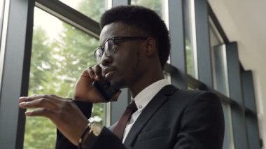 Atış başarılı afro Amerikalı işadamı ile bir takım elbise kadar bina ve cep telefonda konuşurken ofis panoramik pencerelere yakın duran kravat ile kapatın. Telefonda konuşurken memur. Teknoloji