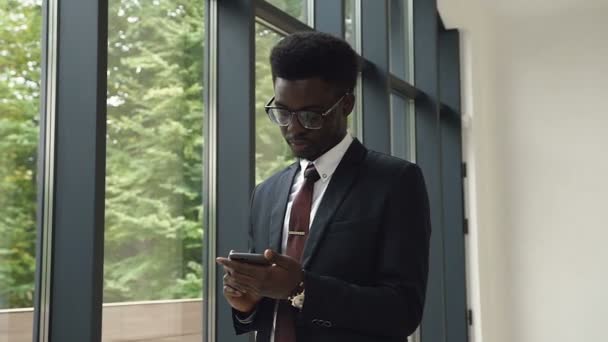 Afrikansk amerikansk mand bruger business app på smartphone, stående i lobbyen på kontor center nær panoramavinduer. Flot ung forretningsmand kommunikerer på smartphone selvsikker – Stock-video
