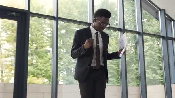 Joven hombre de negocios afroamericano en traje negro está bailando y tira documentos, celebrando el logro de negocios en la oficina con vista a las ventanas panorámicas. De interior — Vídeo de stock