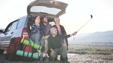Dört selfie smartphone cep telefonu ile yapıyorsun, suv arabanın dağ zemin zemin üzerine eğilerek oturan genç insanlar. Tatil seyahat araba suv, doğa, dağlar, kavramı