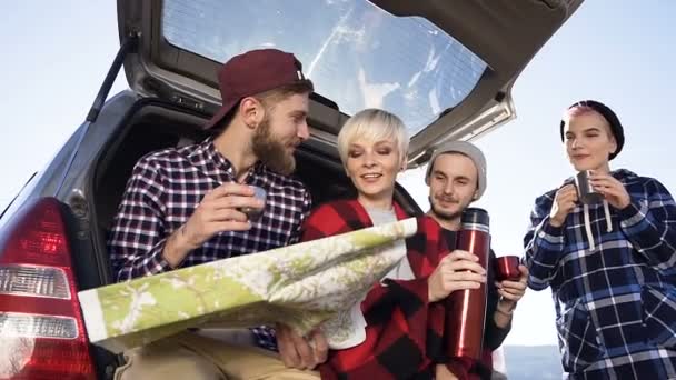 Cuatro turistas alegres tomando café caliente o té miran la hoja de ruta mientras se apoyan en el coche en busca de una nueva ruta de viaje, hablando y riendo. Al aire libre — Vídeo de stock