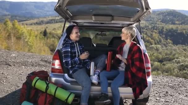Happy friends are leaning on the trunk car drinks coffee or tea on mountains background. Two happy hipster girls sit in the trunk of a car drinking hot coffee or tea from a thermos, talking and — Stock Video