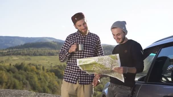 Dos amigos están de pie cerca del auto tomando té mientras miran la hoja de ruta haciendo una ruta más para escalar las montañas, riendo y hablando. Viaja por las montañas en coche. Naturaleza, aire libre — Vídeo de stock