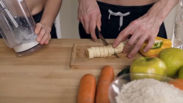 Close up tiro de mãos masculinas cortando banana e mãos femininas misturando leite no misturador na cozinha . — Vídeo de Stock