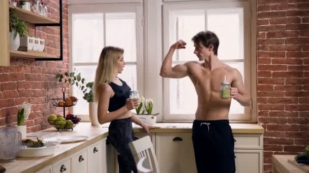 Attractive sport man with smoothie in hands showing his muscles on the arm to beautiful fit woman in the kitchen. — Stock Video