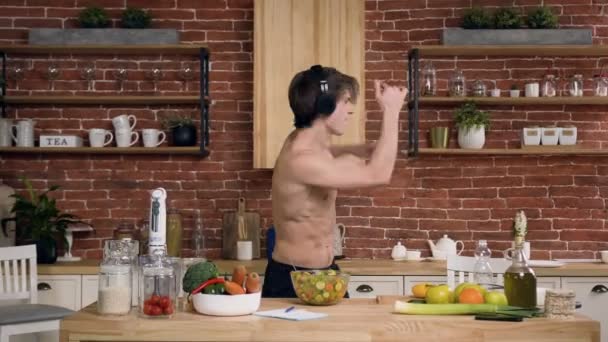 Funny fit hombre escuchando música en los auriculares y bailando mientras cocina . — Vídeos de Stock