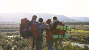 Sırt çantaları ile dört beyaz arkadaş günbatımında dağlar pitoresk bir yerde bekletin. Sarılma, yeşil orman ve nehir üzerinde ileriye doğru bakarak dağların tepesinde duran yolcular