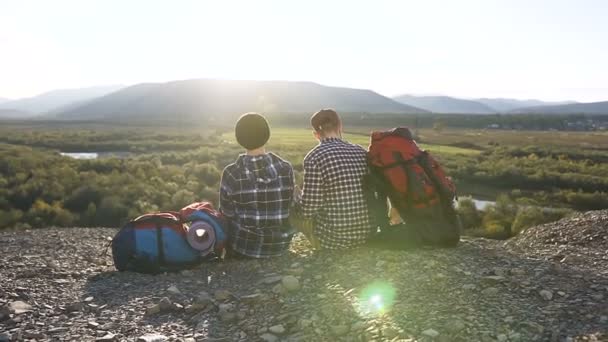 Jovens com uma viagem turística sentam-se no topo da montanha. O menino e a menina nas montanhas admiram as belas vistas das montanhas e da natureza ao pôr do sol . — Vídeo de Stock