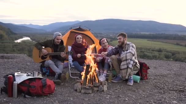 Le jeune voyageur joue de la guitare, ses amis cuisinent des saucisses en feu au coucher du soleil dans les montagnes. Amitié, personnes et musique, concept de voyage. Dolly shot — Video