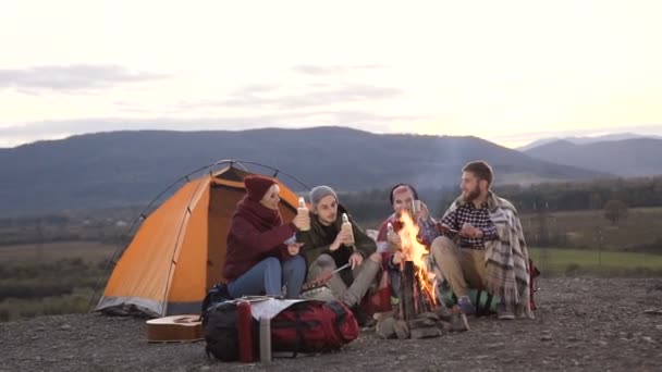 Fyra vita vänner att ha vila nära turist tält med brasa på som stekt korv i bergen. Berg, vandring, ha kul. Fritid, vänskap koncept och turism — Stockvideo