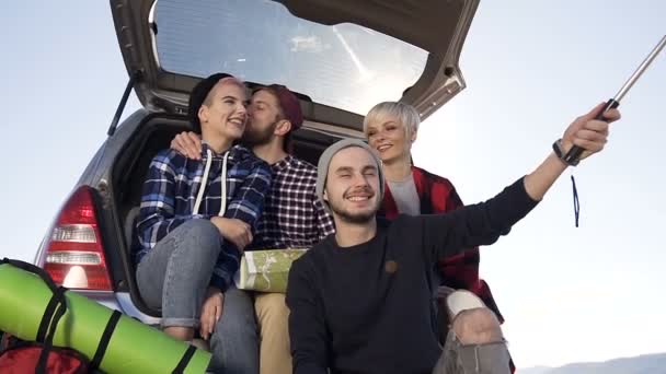 Dos parejas jóvenes haciendo selfie en el teléfono inteligente, sentado en el maletero del coche en el fondo de la montaña . — Vídeo de stock