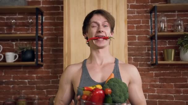 Portrait d'un beau jeune homme souriant avec du piment dans la bouche, debout derrière la table de cuisine et tenant un bol en verre de légumes dans la cuisine moderne à la maison. Concept d'alimentation saine — Video