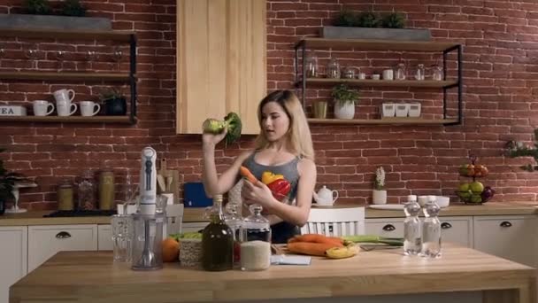 Jeune belle femme vêtue d'un costume de sport debout derrière la table de cuisine avec du brocoli frais à la main, tenant un bol en verre avec des légumes dans la cuisine en face du mur de briques — Video