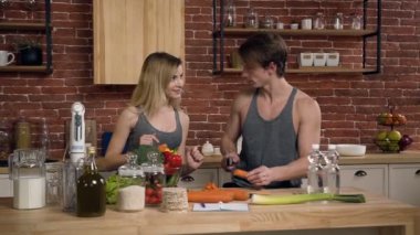 İnsanlar konuşmak ve sağlıklı yemek pişirme sırasında gülümseyen genç spor. Genç adam kesme havuç için taze sebze salata hazırlamak ve kız arkadaşı onu mutfak evde yardımcı olur