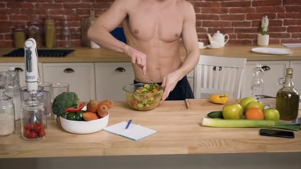 Atractivo chico en forma con torso desnudo escuchando música en los auriculares y bailando mientras cocina ensalada saludable en casa de la cocina. Comida saludable, dieta, cocina — Vídeos de Stock