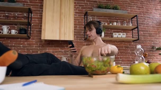 Joven deportista con torso desnudo sentado detrás de la mesa de la cocina comiendo ensalada vegana mientras escucha música en los auriculares y el uso de teléfonos inteligentes para chatear con amigos, sentado en la cocina — Vídeos de Stock