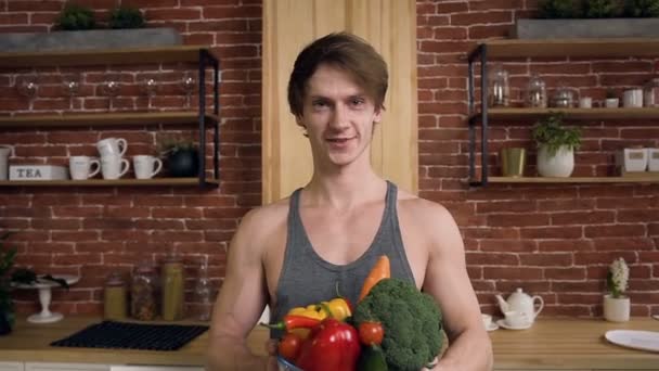 Portrait d'un bel homme musclé souriant tenant un bol en verre avec des légumes frais tout en se tenant à la table de la cuisine avec des aliments sains dans la cuisine moderne. Alimentation saine et mode de vie sain concept — Video