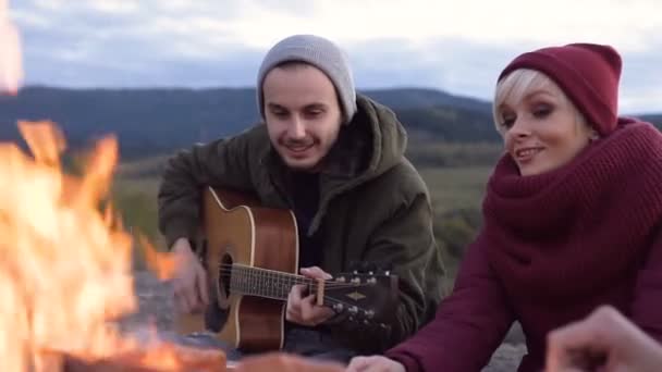 Pohledný mladý pár sedí u ohně a hraje na kytaru na pozadí hory. — Stock video