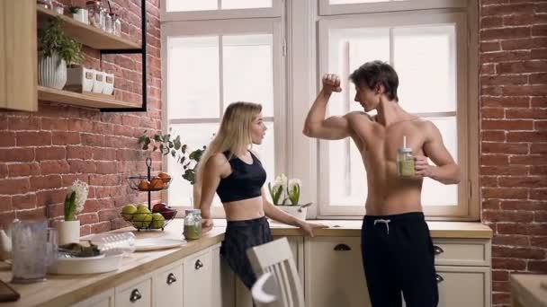 Pareja joven muscular hacer clic con botellas de vidrio beber cóctel saludable después del entrenamiento, hablando y riendo en la cocina. Deporte, dieta, concepto de comida saludable — Vídeo de stock