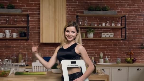 Retrato de una mujer de fitness delgada con báscula eléctrica que promueve la pérdida de peso saludable. Linda mujer en forma en el uniforme deportivo con escamas en la mano y posando en la cámara en el fondo de la cocina — Vídeos de Stock