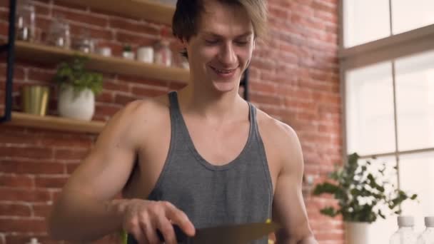 Guapo joven sonriente está cortando pimentón amarillo y tratando pone trozos picados en un tazón de vidrio para preparar ensalada fresca en la cocina. Dieta, concepto de comida saludable. Dolly disparó — Vídeo de stock