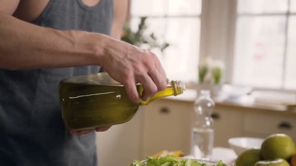 Close-up. Jovem é derrama de garrafa um azeite em tigela de vidro com legumes para preparar salada fresca na cozinha doméstica. Estilo de vida saudável, nutrição adequada, pratos veganos — Vídeo de Stock