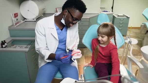 African dentist teaching little female patient how to brush teeth on the plastic model. — Stock Video