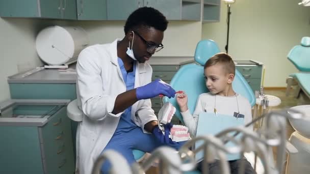 Afrikanska unga tandläkare lära liten pojke att borsta tänder på plast modell. — Stockvideo