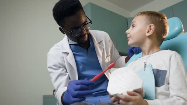 Молодой африканский дантист учит маленького мальчика пользоваться зубной щеткой пластиковыми зубами . — стоковое видео
