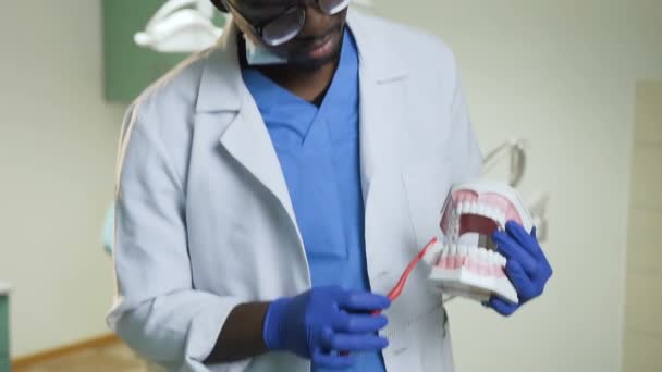 Dentista africano que muestra cómo limpiar los dientes con cepillo de dientes en un modelo de plástico . — Vídeo de stock