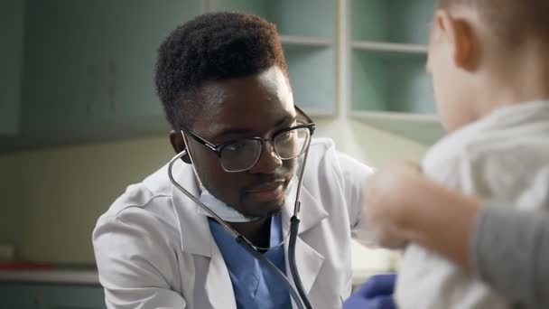 Joven pediatra africano examinando a un pequeño paciente masculino usando estetoscopio en la oficina del hospital . — Vídeos de Stock