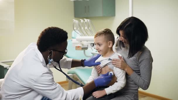 Joven pediatra africano revisando pulmón de niño pequeño usando estetoscopio en la oficina . — Vídeos de Stock
