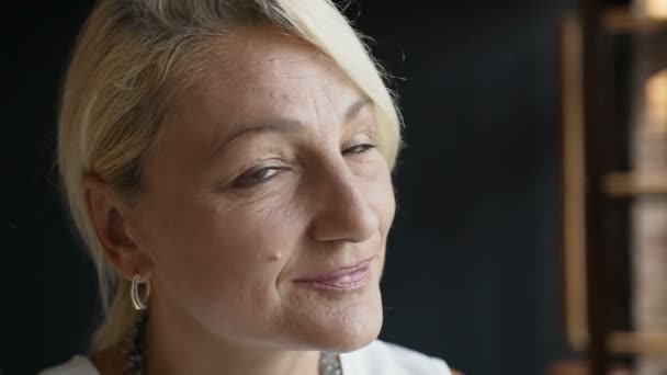 Portrait of the stylish middle aged blonde is businesswoman in stylish clothes smiling pensive at camera indoors on the background dark wall — Stock Video